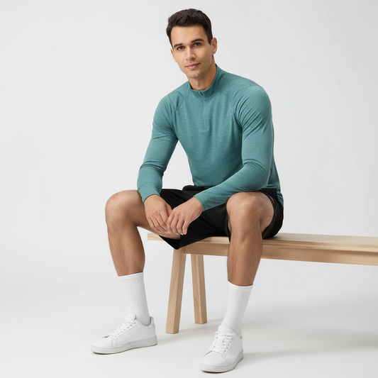 Deniz yeşili erkek uzun kollu quarter zip yüksek yaka antrenman üstü, beyaz fonda oturan model üzerinde; nefes alan ve esnek performans kumaş.